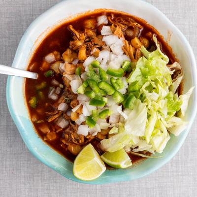 Red chile broth soup with hominy, shredded meat, lettuce, onion, and lim.