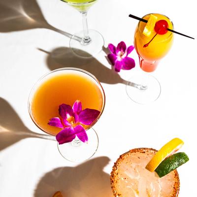 Four colorful cocktails garnished with fruit and flowers cast shadows on a white table