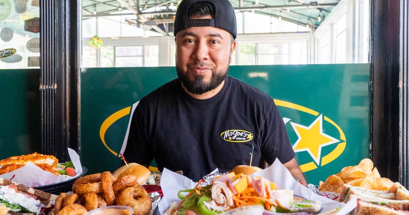 Chef  in a black cap and shirt sits at a table overflowing with food