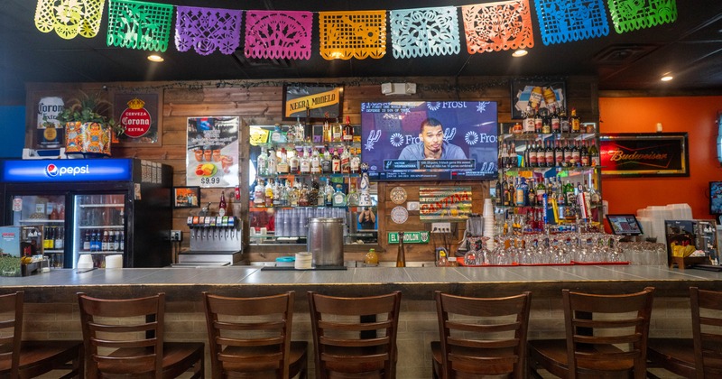 Interior of a bar with colorful papel picado banners, wooden bar counter, chairs, liquor bottles