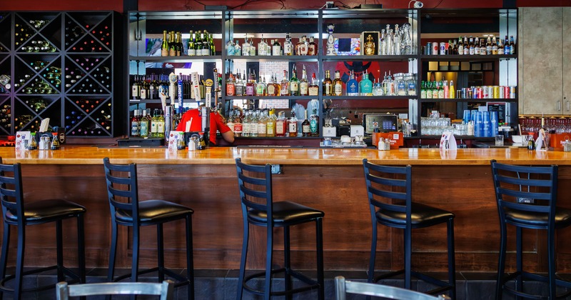 A bar with a wooden counter and high-backed stools, shelves stocked with liquor bottles behind.