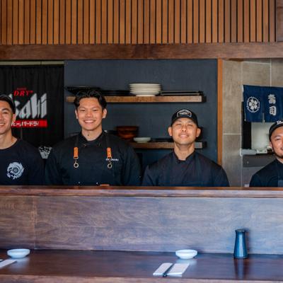Sushi chefs standing behind a wooden counter inside a modern Japanese restaurant.