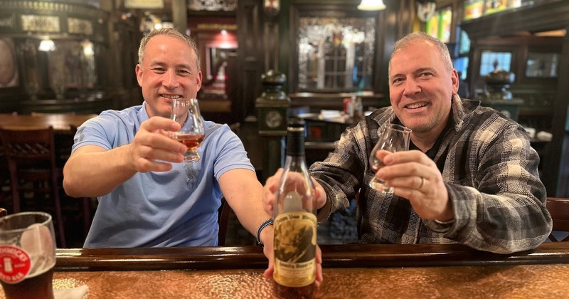 Two patrons smiling and raising glasses of whiskey at a cozy bar