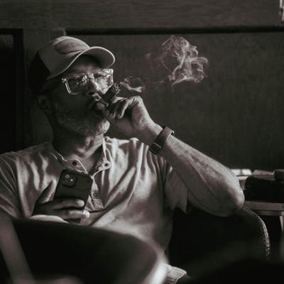 A patron in a cap and glasses smokes a cigar, holding a phone, with a relaxed expression.