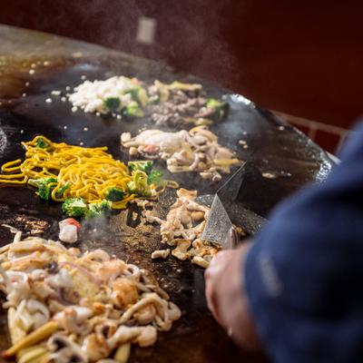 A chef cooks noodles, meats, and vegetables on a large round flat-top grill.