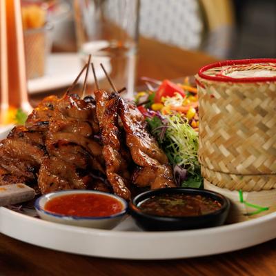 Thai grilled pork skewers served with dipping sauces, rice in a small basket, and salad.