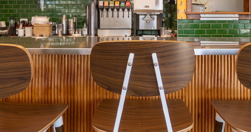 An interior view - high wooden stools, a counter, green tile wall, menu boards and drink dispensers