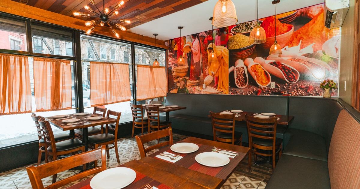 Dining area with wooden chairs and tables set with plates; large windows with orange curtains.