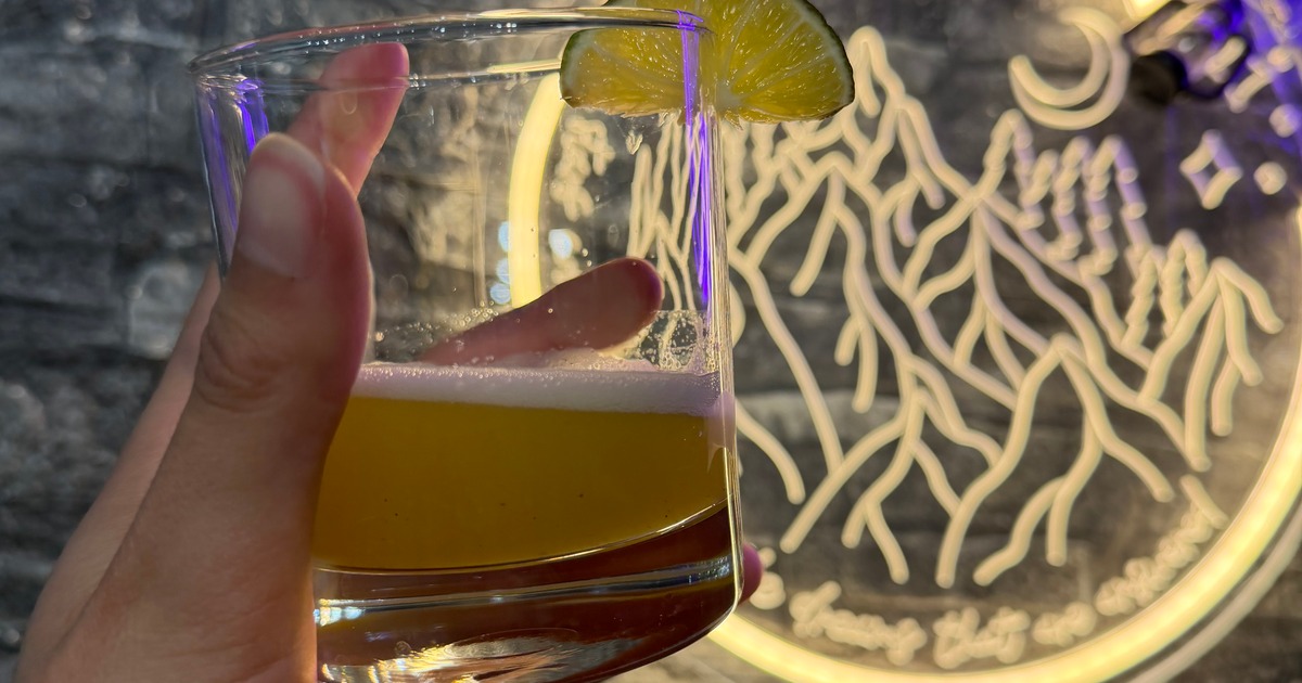 Hand holding a yellow cocktail with lime garnish before a mountain neon sign