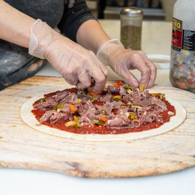 Hands in gloves topping tomato-based pizza dough with meat and vegetables.