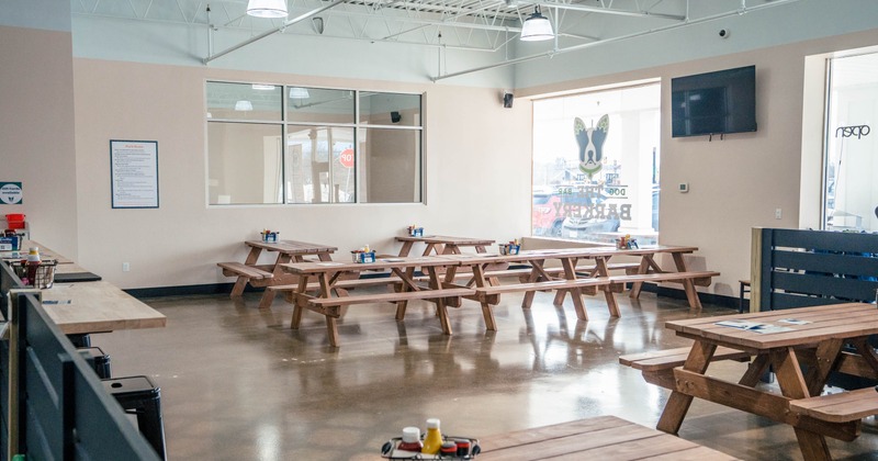 Interior of a room with wooden picnic tables, large windows, and industrial lighting