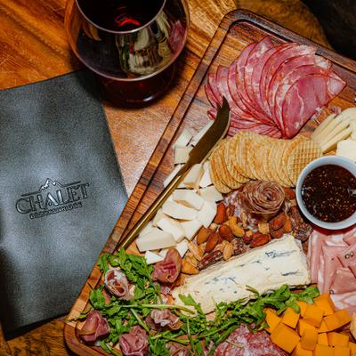 A charcuterie board sits beside a glass of red wine and a menu on a wooden table..