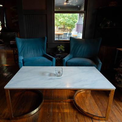 A cozy seating area with two teal armchairs and a marble coffee table in front of a window.
