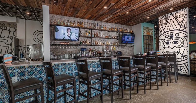 A modern bar with a rustic wooden ceiling, vibrant patterned tiles, and nine black chairs