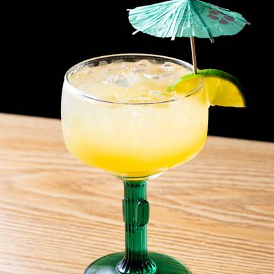 Yellow colored Margarita served in a glass with cactus shaped stem, lime, cocktail umbrella.