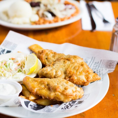 Fried fish pieces on top of chip style french fresh next to a side of coleslaw, lemon and tartar sauce