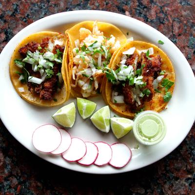 Trio of traditional Mexican tacos with a dipping sauce and garnishes.