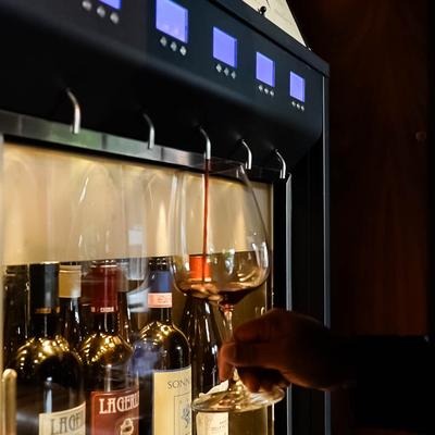 A wine dispenser pours red wine into a glass held by a person, with displayed wine bottles.