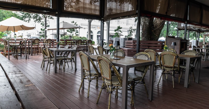Covered outdoor restaurant patio with woven chairs, tables, umbrellas, and retractable shades