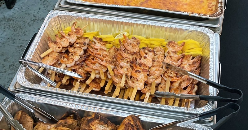 A catering setup featuring separate serving trays with shrimp skewers, chicken, and other dishes