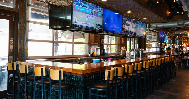 A bar - a long wooden counter, black stools, multiple TVs, a colorful surfboard decoration.