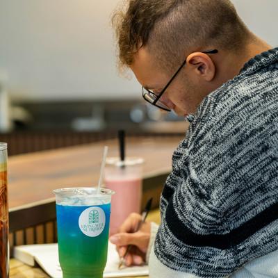 Person writing in a notebook at a counter while having a drink.