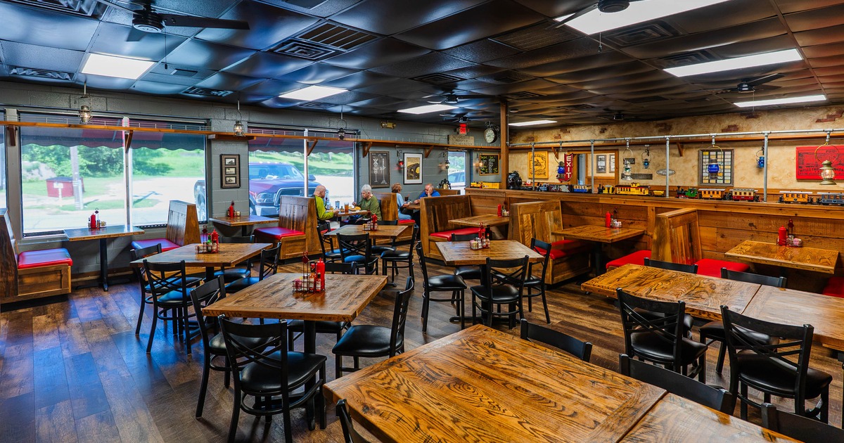 Interior of a rustic restaurant with wooden tables and black chairs, featuring large windows