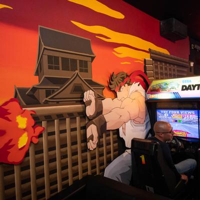 A person plays an arcade racing game next to a vibrant mural of Street Fighter's Ryu character.