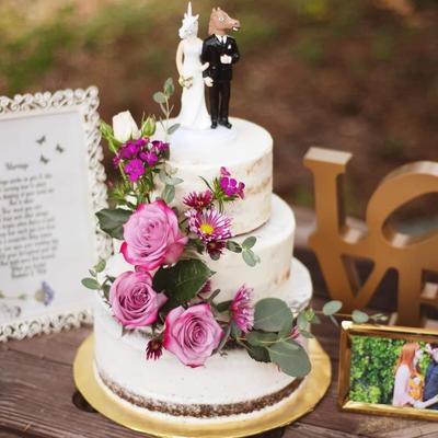 A cake topped with humorous unicorn and horse figurines, nearby, a framed message and a picture.