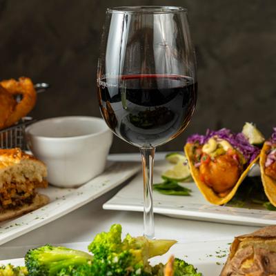 A glass of red wine surrounded by plates of fish tacos and broccoli.