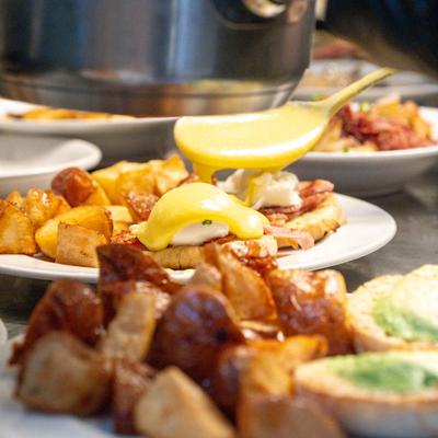 Eggs Benedict being drizzled with hollandaise sauce, surrounded by roasted potatoes.
