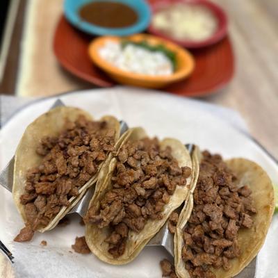 Tacos filled with finely chopped meat, served with dishes of salsa and toppings in the background.
