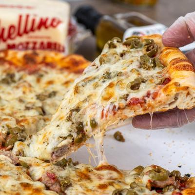 A hand lifting a slice of pizza with melted cheese and green olives.