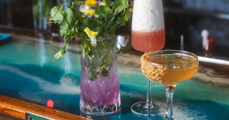 Three colorful cocktails on a blue glass bar counter with a Tequila Way sign