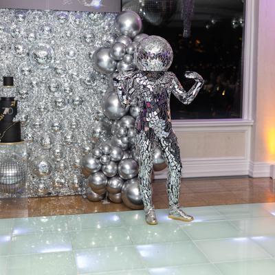 Mirror covered performer posing beside silver balloon display.