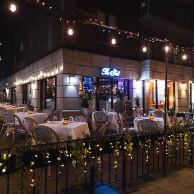 A night scene of a restaurant with outdoor dining, featuring set tables and string light decoration.