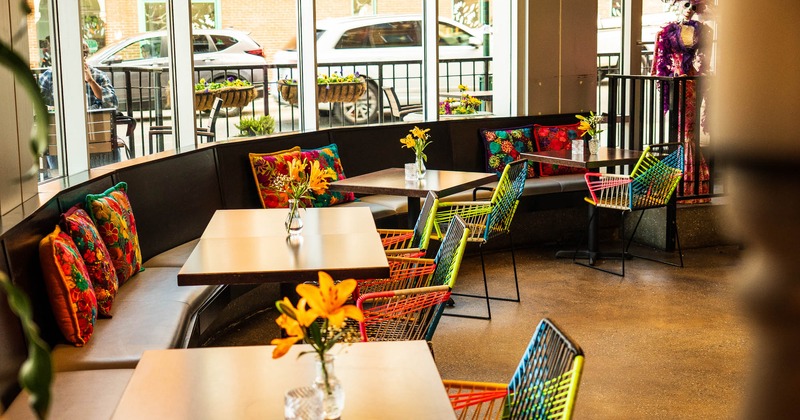 Long banquette seating by a window side, with colorful pillows and chairs, flowers on tables