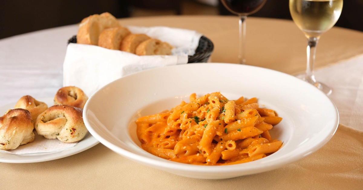 Penne ala vodka pasta plate sits on a table with garlic knots, bread, and served wine