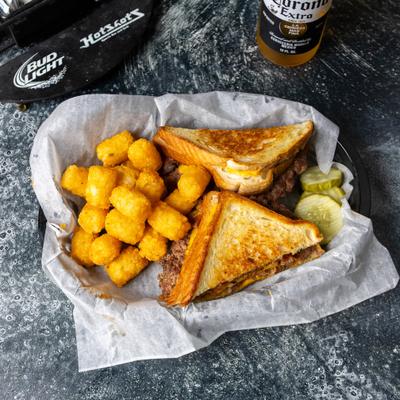A basket containing tater tots, a patty melt sandwich, and sliced pickles.