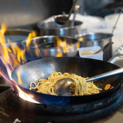 Noodles in a flaming wok, pots in the background.