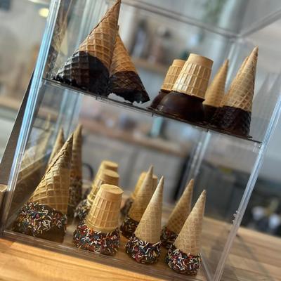 Waffle cones in a display case, some dipped in chocolate with rainbow sprinkles or plain.
