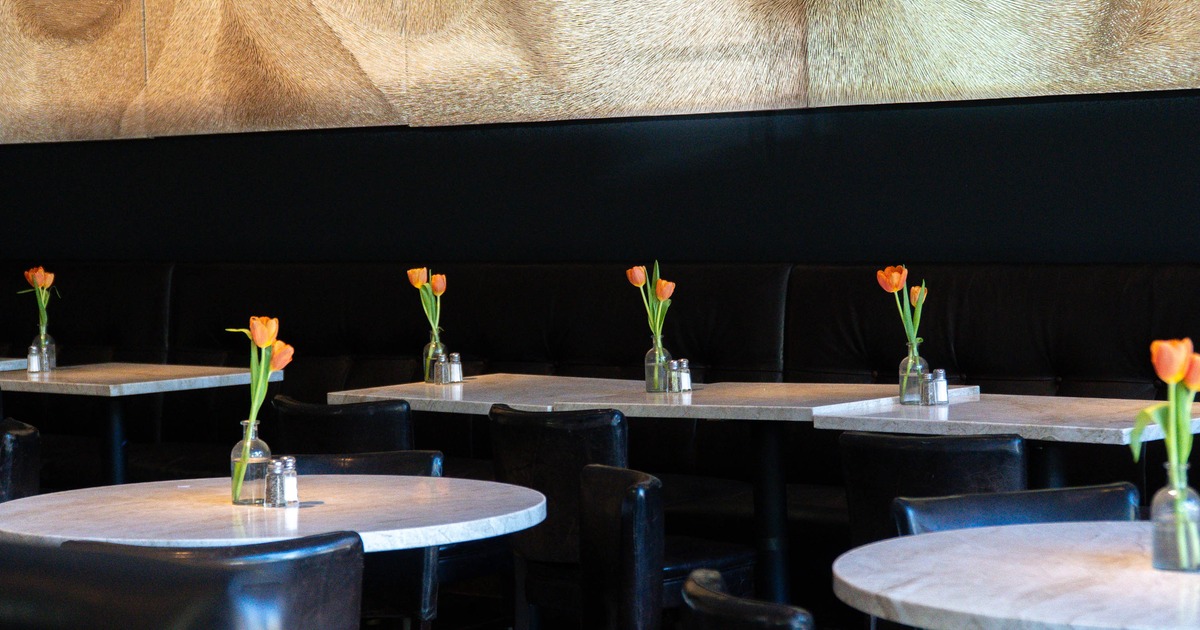 Restaurant interior with round tables, black seating, orange tulips in glass vases, and large abstract wall art
