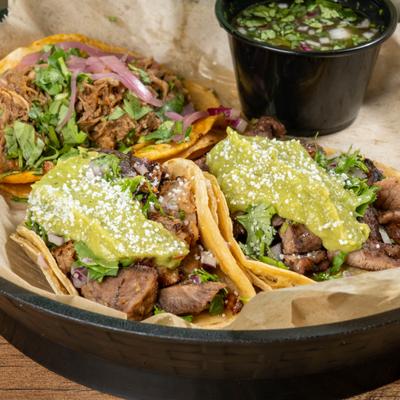 3 meat tacos with guacamole and cotija on a plate, side of sauce.