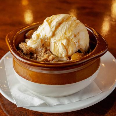 Apple cobbler topped with melting vanilla ice cream in a brown ceramic bowl on a white plate.