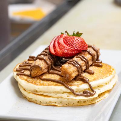 Pancakes topped with two churros, chocolate drizzle, and a sliced strawberry.