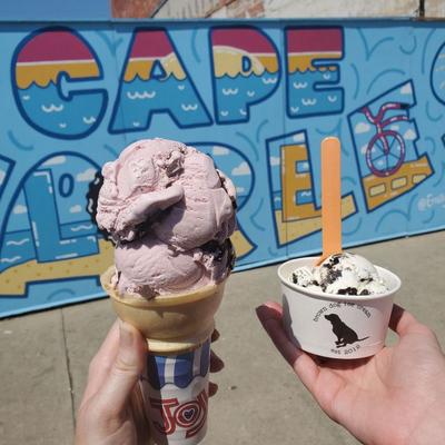 Cone with pink ice cream and a cup with white ice cream, held in front of a colorful mural.