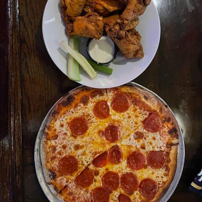 A pepperoni pizza and a plate of chicken wings with celery sticks and a dipping sauce.