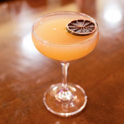 An orange cocktail served in a coupe glass with dry citrus wheel.