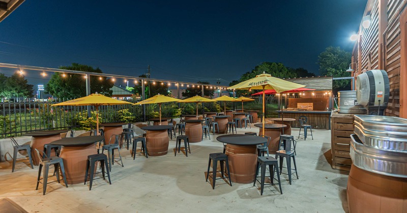 Outdoor seating area, barrel tables, metal stools, and yellow umbrellas under string lights