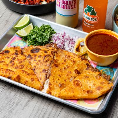 Quesabirria slices served consommé, chopped onions, cilantro, and lime wedges.